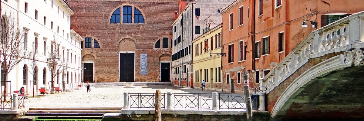 Colour photo: view of the square and the façade of the church of the same name, built in red brick. View from the opposite bank of the canal, whose water can be seen in the foreground with the bridge on the right. On the right-hand side is a group of houses in various colours, and on the left-hand side is a group of white houses. The square is in the middle, with the church in the background.