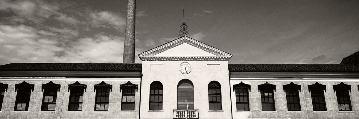 Black and white photograph: depicts the main façade of the spinning mill, in Palladian villa style, with a gate with rounded iron railings at the top. At the centre of the upper part is a round white stone clock, in front of the spinning mill, grass and land. Behind it is a clear sky with a few white clouds on the left and the red brick chimney of the furnace.