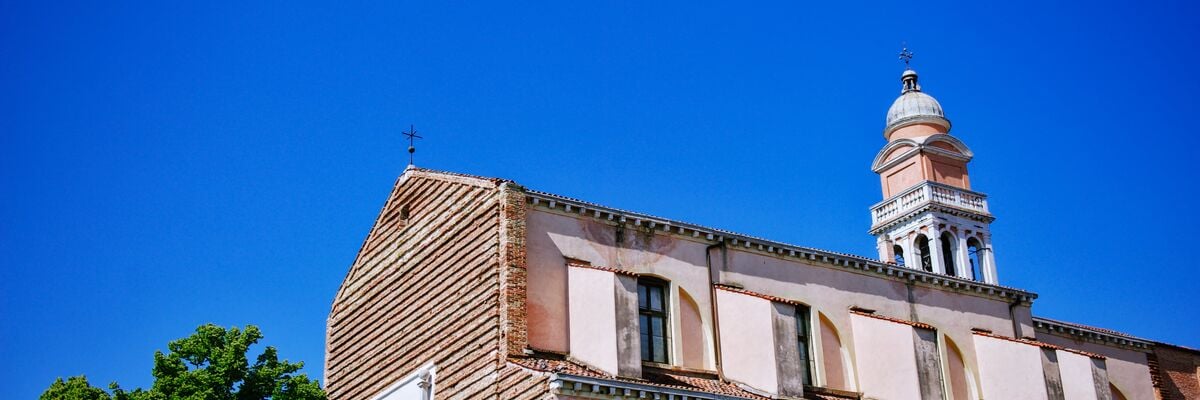 Colour photograph: the upper corner of a historic building made of pink stone with a tiled roof, framed against a deep blue sky. The building features arched windows, a small bell tower with a dome, and a cross. Part of a green tree can be seen on the left.