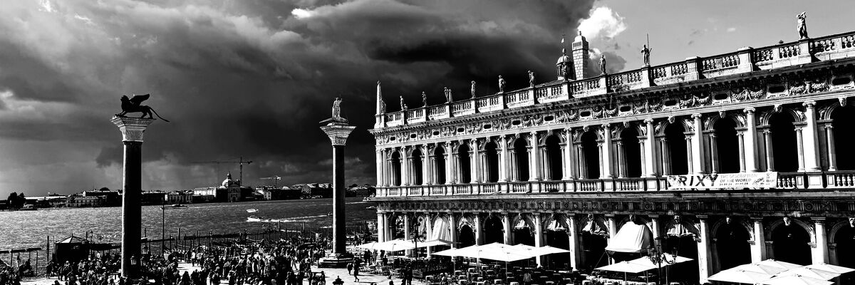 Foto in bianco e nero: le colonne di San Marco e Todaro, la Biblioteca Nazionale Marciana, il bacino di San Marco e molta gente a passeggio. Nuvoloni scuri all'orizzonte.