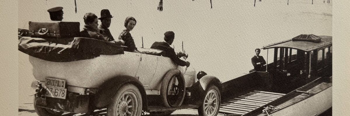 Vintage black-and-white photograph showing a car with five passengers being loaded onto a makeshift ferry boat at the Lido of Venice, with the lagoon and distant buildings in the background.