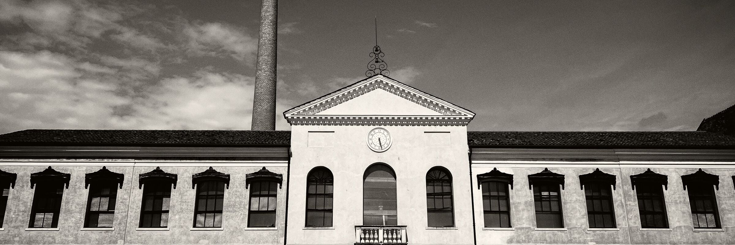 Black and white photograph: depicts the main façade of the spinning mill, in Palladian villa style, with a gate with rounded iron railings at the top. At the centre of the upper part is a round white stone clock, in front of the spinning mill, grass and land. Behind it is a clear sky with a few white clouds on the left and the red brick chimney of the furnace.