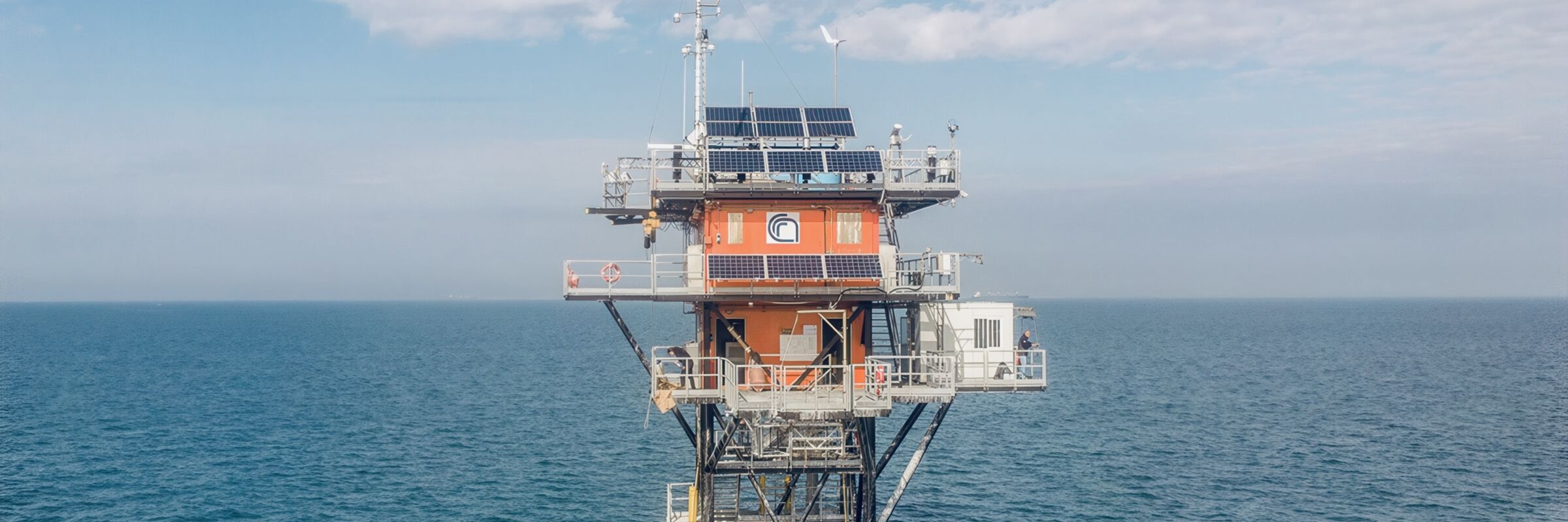Foto a colori della torre oceanografica fotografata in volo. Sotto e intorno il mare e un cielo azzurro con nuvole bianche. La torre è un'infrastruttura in tubi d'acciaio, con due unità abitative di colore arancione, un box esterno bianco finestrato e vari sensori distribuiti in tutta la struttura.